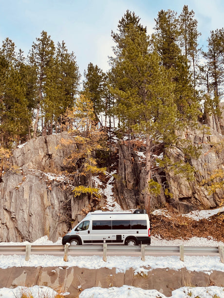 An RV rental, a silver camper van, is parked on a snowy road beside a towering rock face and evergreen trees. Ideal for winter adventures.