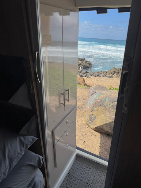 From an RV rental, a cozy interior with bed and cabinets overlooks a sunny beach, ocean waves, and rocky shore with distant people.