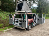 This customized RV rental camper van with a pop-top tent and open doors showing its kitchen setup is parked in a forest.