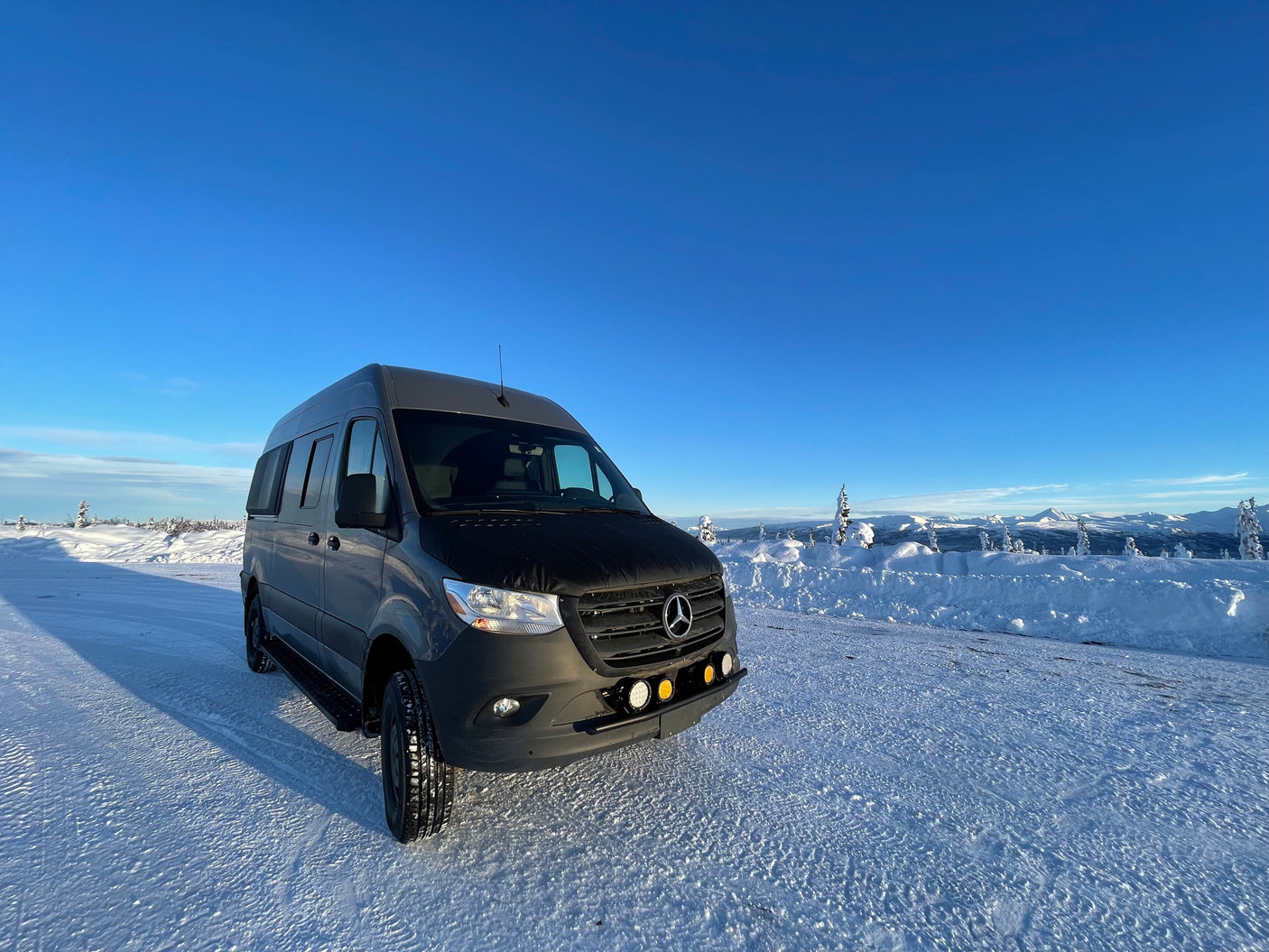 Dark grey Mercedes Sprinter rv rental van on a snowy road, flanked by mountains under a clear blue sky.
