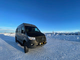Dark grey Mercedes Sprinter rv rental van on a snowy road, flanked by mountains under a clear blue sky.