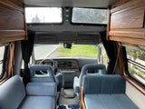 Inside an rv rental: view from the back, showing blue upholstered seats, wooden cabinets, and the cockpit with road view.
