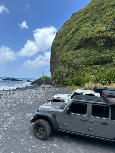Jeep Gladiators on a black sand beach by a lush cliff and ocean. Great for an adventure rv rental exploring scenic coasts.