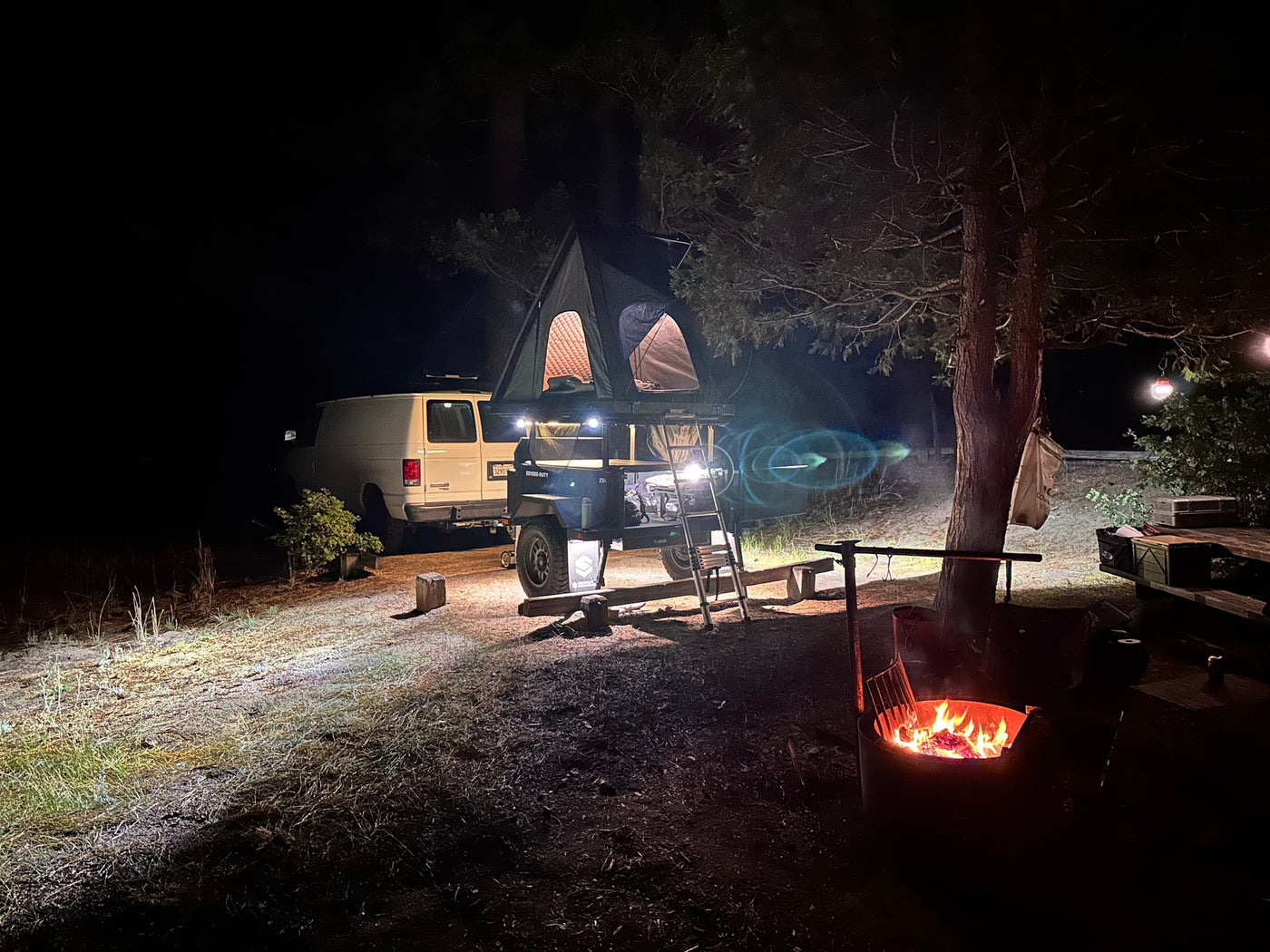 Cozy night camping: a white van, camper trailer with a rooftop tent, and glowing campfire under trees. An RV rental experience. — photo 6