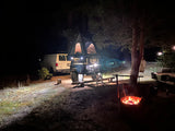 Cozy night camping: a white van, camper trailer with a rooftop tent, and glowing campfire under trees. An RV rental experience.