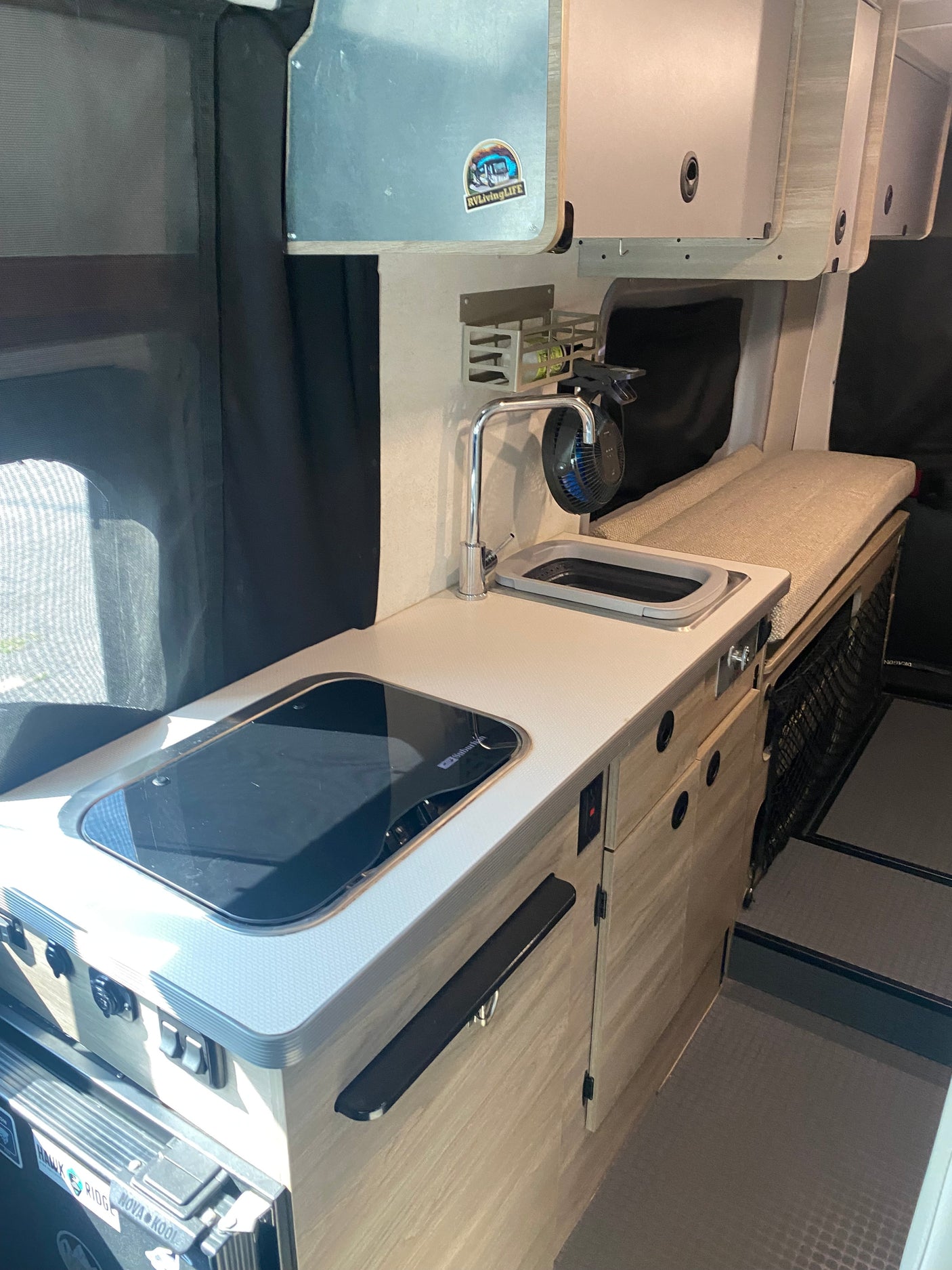 RV rental interior: compact campervan kitchenette with a stovetop, sink, faucet, and overhead cabinets, alongside a cushioned bench.