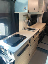 RV rental interior: compact campervan kitchenette with a stovetop, sink, faucet, and overhead cabinets, alongside a cushioned bench.