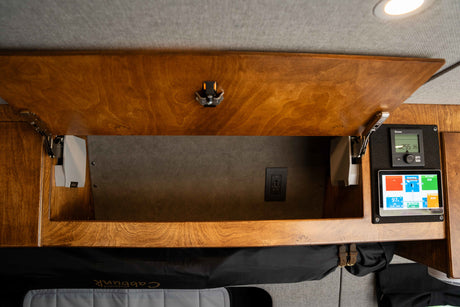 Inside an RV rental: open overhead wood cabinet, power outlet, and energy monitors. Practical amenities for van life.