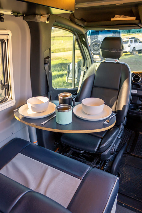 Inside a modern RV rental, a swivel table is set for two with bowls, plates, and mugs for a cozy meal.