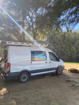 A white Ford Transit camper van parked on a sandy shore by trees, bathed in sunlight. An inviting RV rental for nature trips.