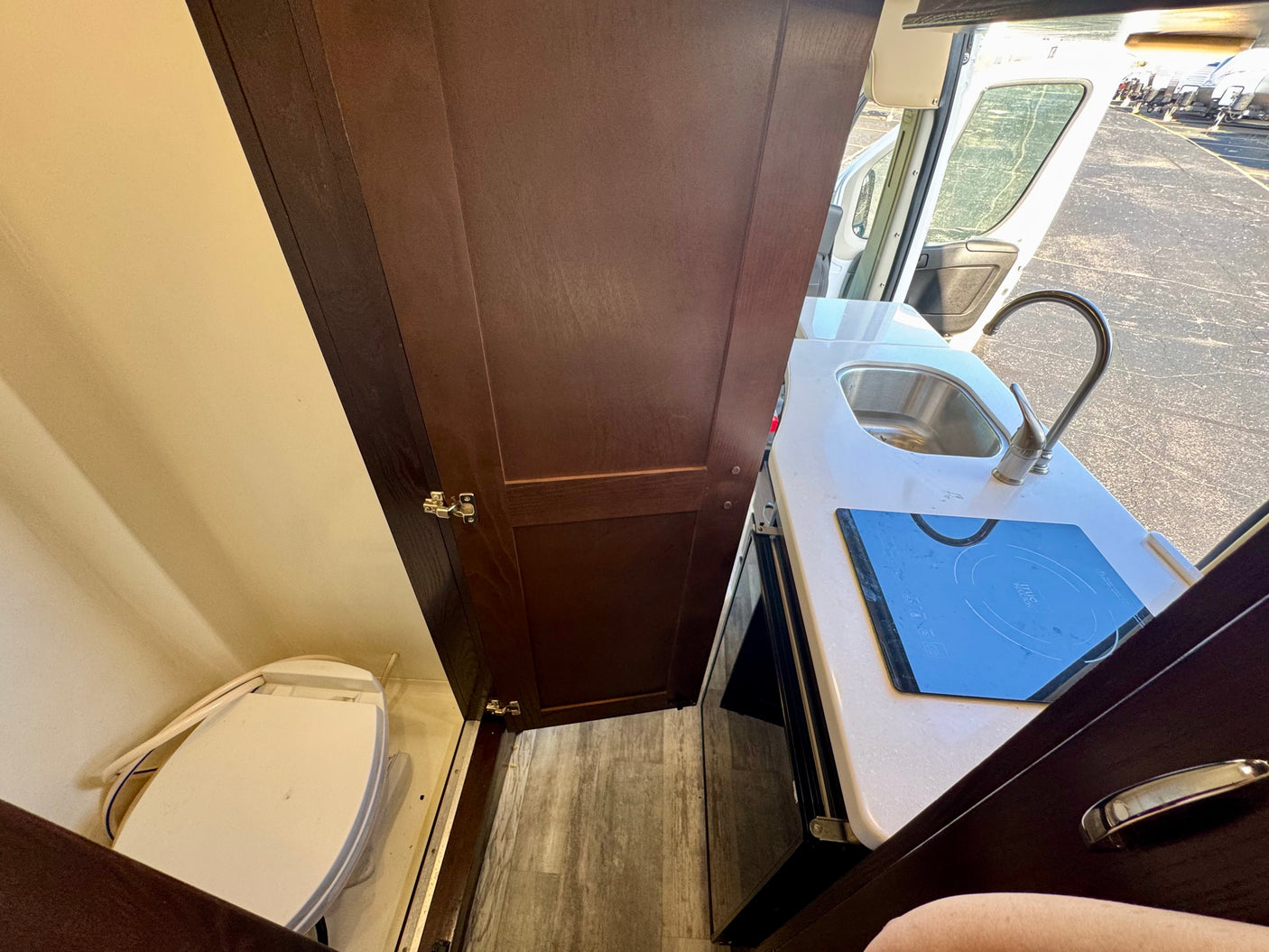 Compact RV rental interior featuring a toilet, dark wood cabinetry, and a small kitchen with a sink and induction cooktop.