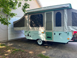 Charming light blue pop-up RV rental with nature decals. Parked outside a garage, ready for your trip.