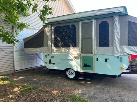 Charming light blue pop-up RV rental with nature decals. Parked outside a garage, ready for your trip.