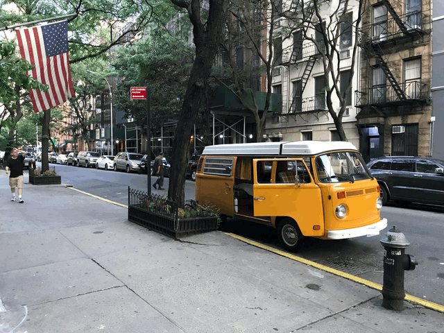 A bright yellow vintage VW camper van, possibly an RV rental, is parked curbside on a city street with an American flag.