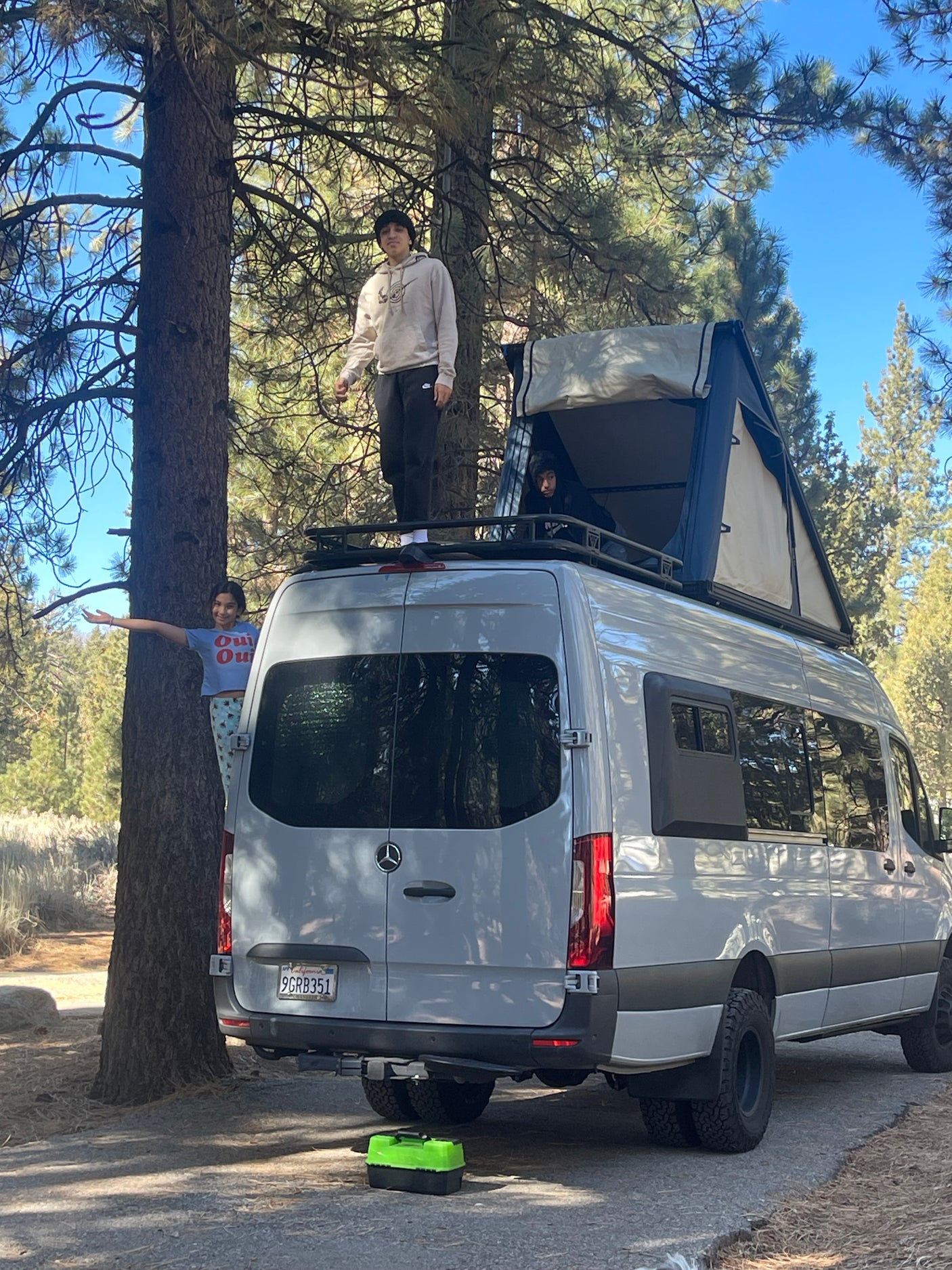 Enjoying the outdoors with an RV rental! Kids explore a Mercedes Sprinter van, one on the roof rack, another in the rooftop tent, third by tree.