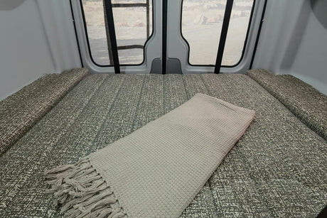Experience an RV rental! This camper van's cozy bed, covered with a waffle-knit blanket, offers views from the rear doors.