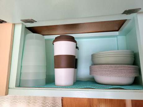 RV rental kitchen cabinet: Organized compact storage with stacked cups, travel mugs, bowls, and paper plates.
