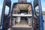 Rear view of an RV rental with open doors, revealing a spacious camper van interior featuring a bed, kitchen, & storage.