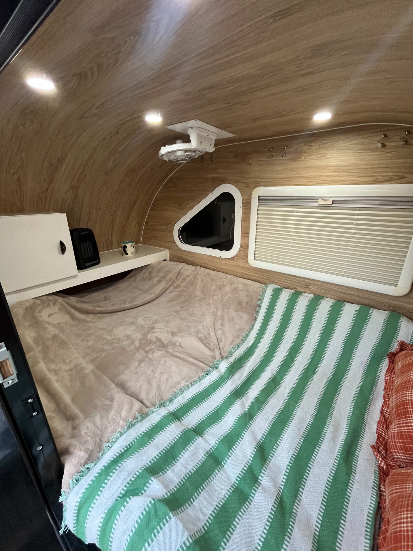 Cozy RV rental interior features a comfortable bed with a green blanket, wood-paneled ceiling, shelf, and windows.