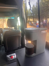 RV rental interior: Coffee maker on counter, fire extinguisher near seat. Overlooking a lively campsite, ready for adventure.
