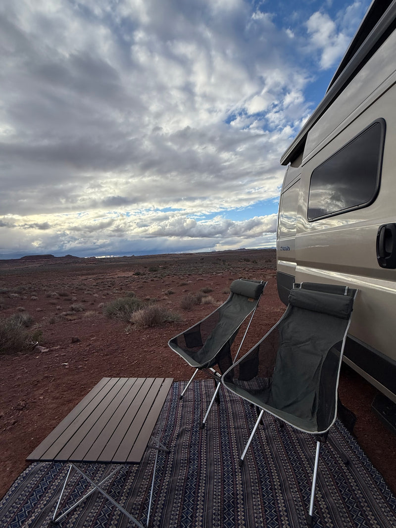RV rental set up with camping chairs and table on a mat in a scenic desert landscape under a dynamic, cloudy sky. — photo 5
