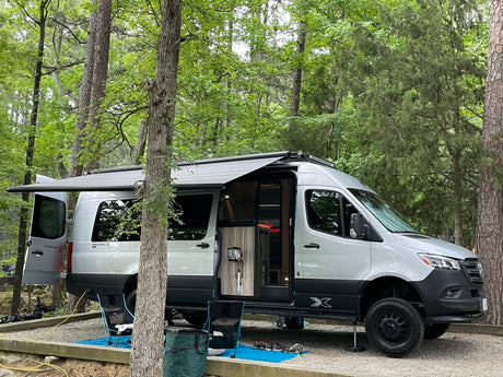This Mercedes-Benz Sprinter camper van, possibly an RV rental, is set up at a wooded campsite with its awning extended and doors open, ready for outdoor living.