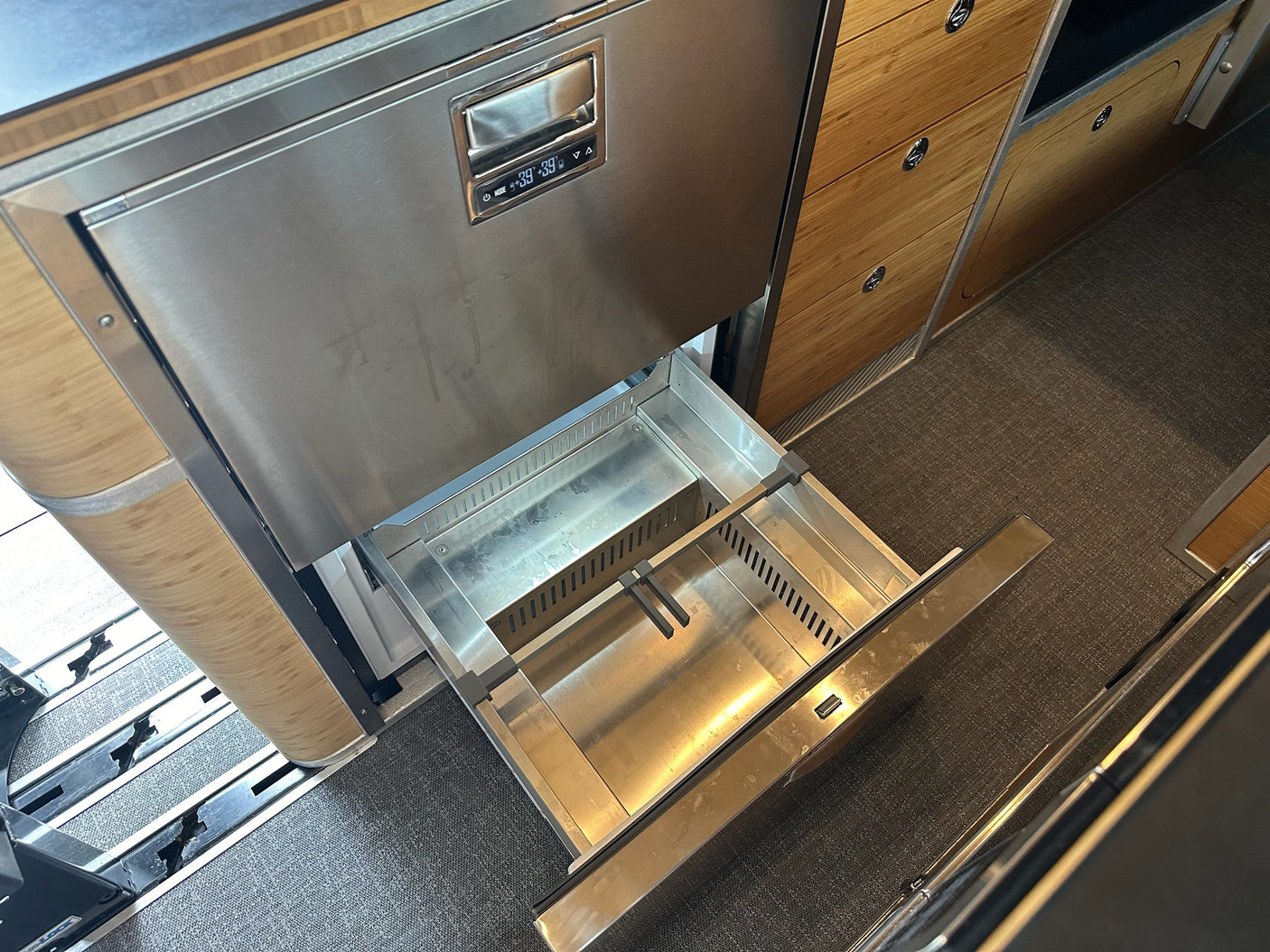 Inside an RV rental: Open stainless steel pull-out fridge/freezer drawer, showing 39°F, by wood cabinets.