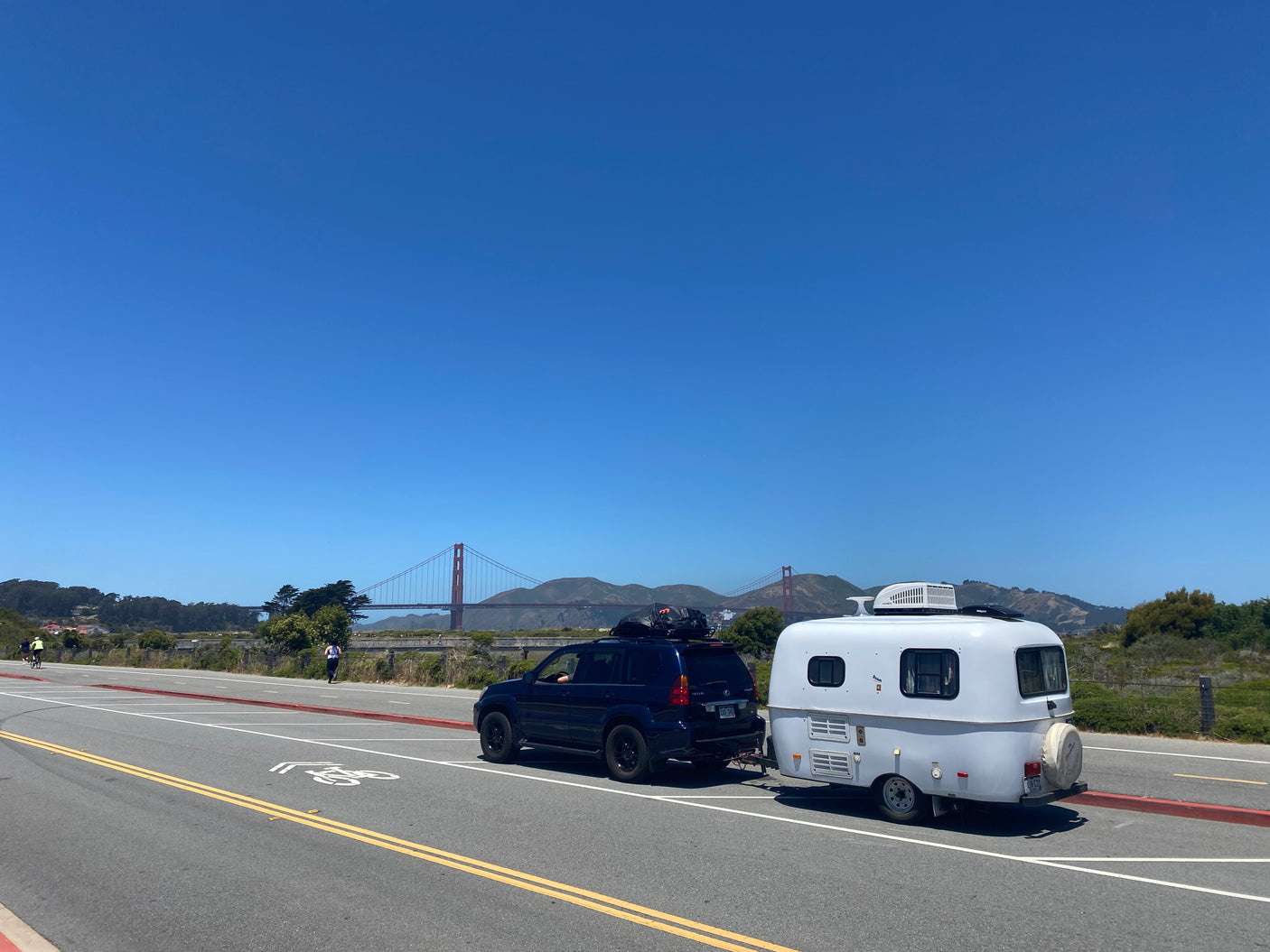 RV rental trip: a compact white travel trailer towed by an SUV, parked near the Golden Gate Bridge under a clear sky.
