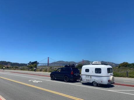 RV rental trip: a compact white travel trailer towed by an SUV, parked near the Golden Gate Bridge under a clear sky.