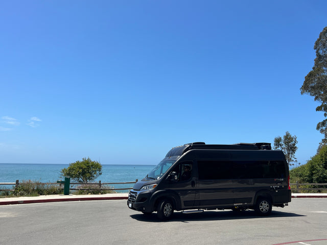 Dark grey Ethos RV rental van parked by the ocean under a clear blue sky, ready for coastal adventures.