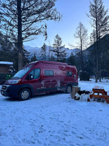 A red RV rental parked in a snowy mountain campsite, with a dog sniffing the ground by a picnic table.