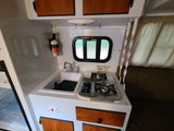 Compact RV rental kitchen featuring a sink, two-burner stove, fire extinguisher, overhead cabinets, and a small window.
