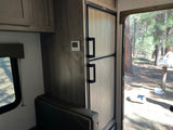 Inside an RV rental: a Dometic fridge, thermostat, and a sunlit forest campsite view through the open door.