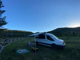 A modern RV rental van with an awning, table, & blanket set up for a peaceful campout in a grassy mountain valley.