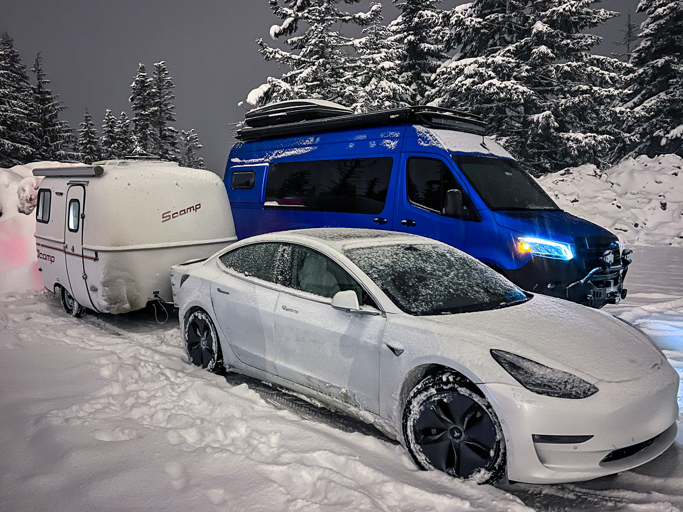 Snow-covered Tesla towing a Scamp camper, parked next to a blue van, ready for a winter rv rental trip.