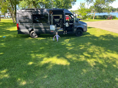 A family enjoys their RV rental camper van parked on a sunny lakeside campsite, with children playing outside.