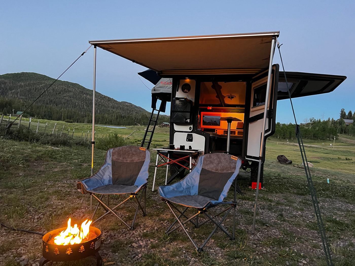Enjoying an RV rental overland trailer with a warm fire pit and chairs at dusk, showcasing its illuminated interior against mountains. — photo 13