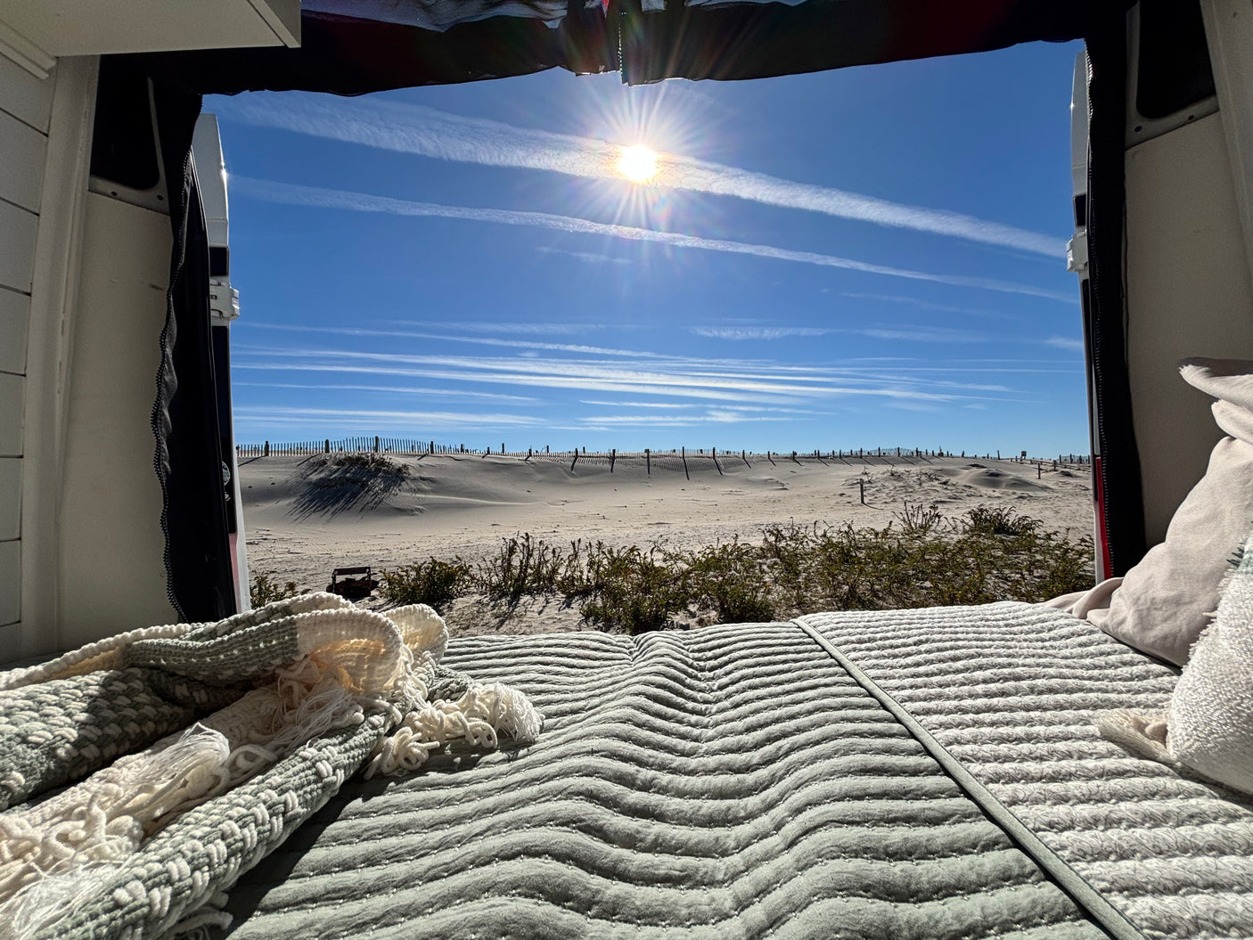 RV rental comfort: a cozy bed with a scenic view of sunny dunes, a beach fence, and a brilliant blue sky.