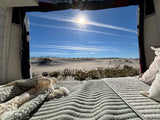 RV rental comfort: a cozy bed with a scenic view of sunny dunes, a beach fence, and a brilliant blue sky.