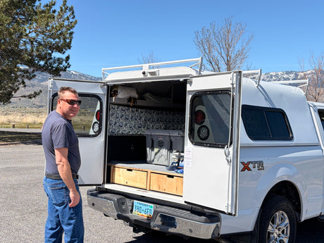 Smiling man by his white truck camper, doors open to custom storage & sleeping platform. Ideal for overlanding; a compact rv rental idea.