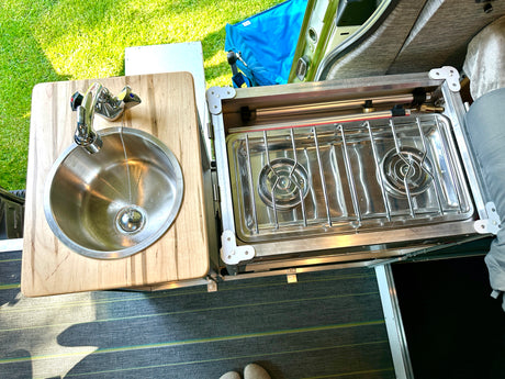An rv rental's outdoor kitchen setup: wooden counter with sink and a two-burner stove, visible with green grass outside.
