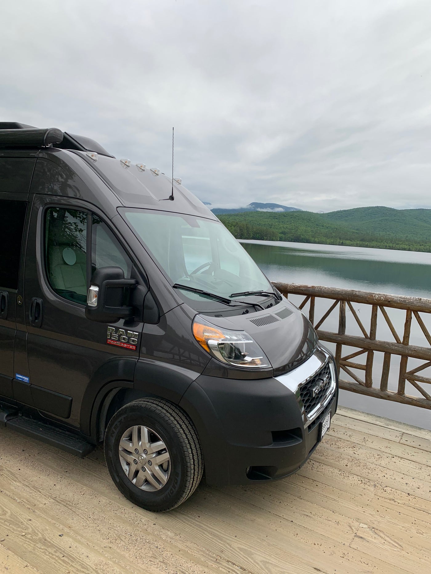 Scenic RV rental adventure: a dark grey Ram ProMaster camper van on a wooden dock by a calm lake with forested mountains. — photo 3
