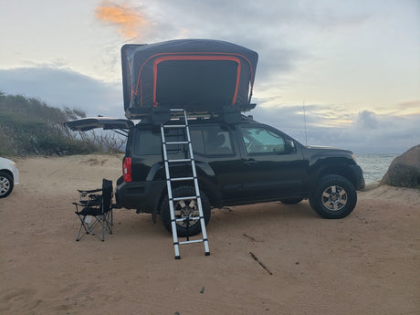 RV rental adventure: Black SUV with rooftop tent & ladder, camped on a sandy beach at dusk.