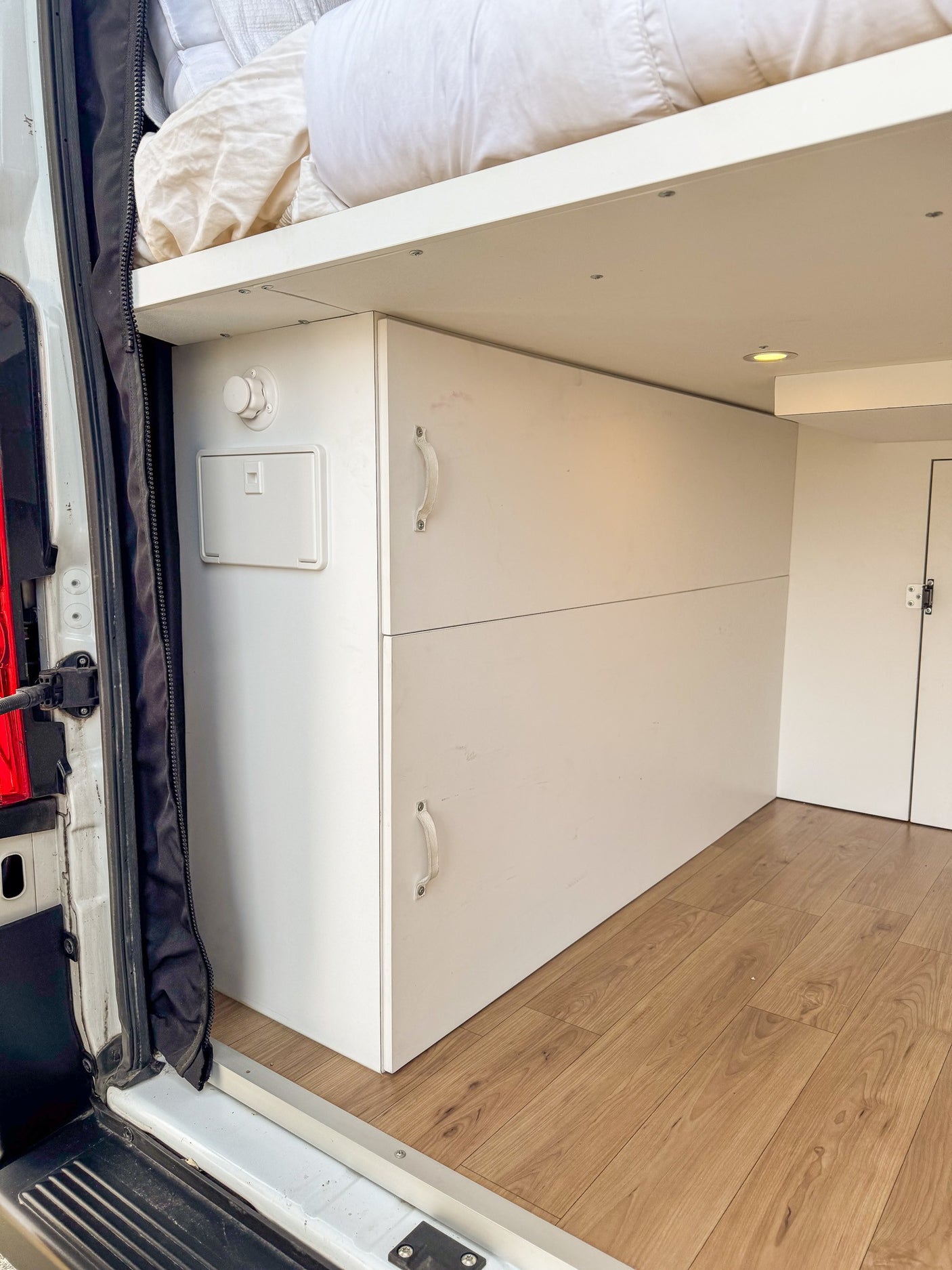 Bright rv rental camper van interior: elevated bed, white storage, wood-look floor, and utility panel by the door.