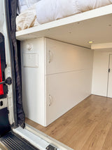 Bright rv rental camper van interior: elevated bed, white storage, wood-look floor, and utility panel by the door.