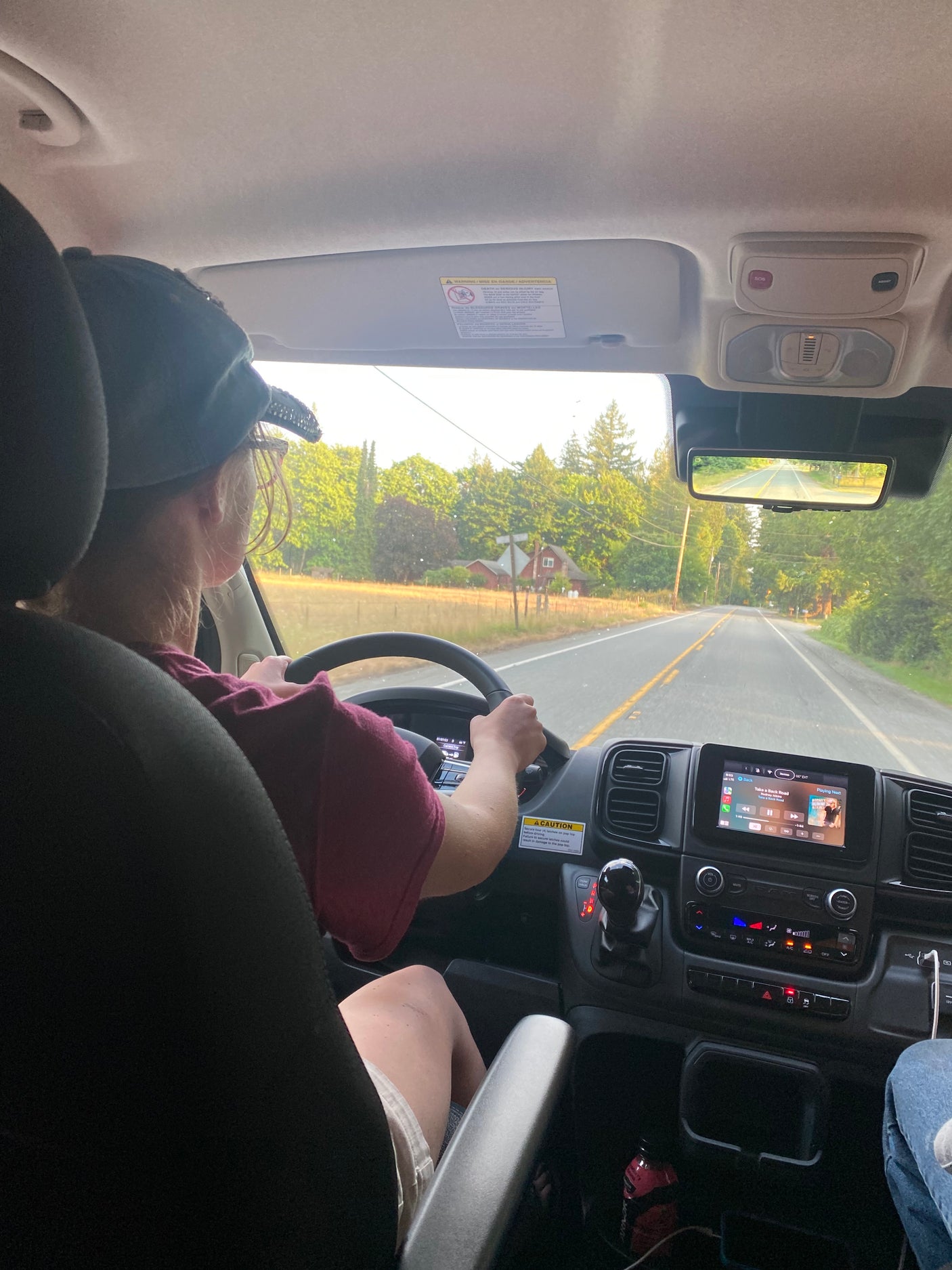 Inside an RV rental, a driver navigates a tree-lined road. The dashboard and infotainment screen are clearly visible.