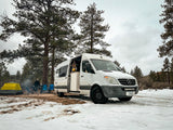 Winter camping with an RV rental van in a snowy pine forest. A person by a campfire and tent complete the cozy van life scene.