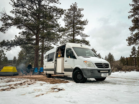Winter camping with an RV rental van in a snowy pine forest. A person by a campfire and tent complete the cozy van life scene.