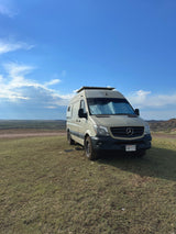 RV rental: A Mercedes Sprinter campervan with solar panels is parked on a grassy hill overlooking a scenic landscape under a cloudy sky.