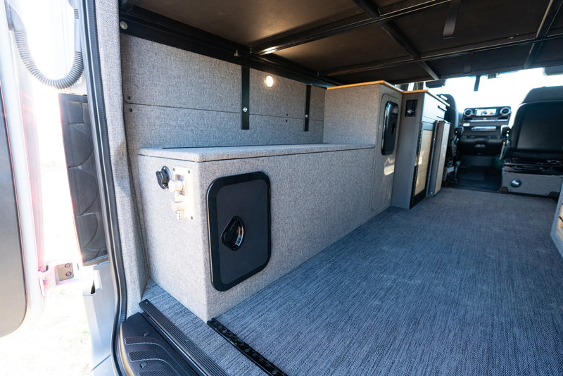 Inside this rv rental, a functional rear area features grey fabric walls, storage boxes, carpeted floor, and an overhead bed platform. — photo 14